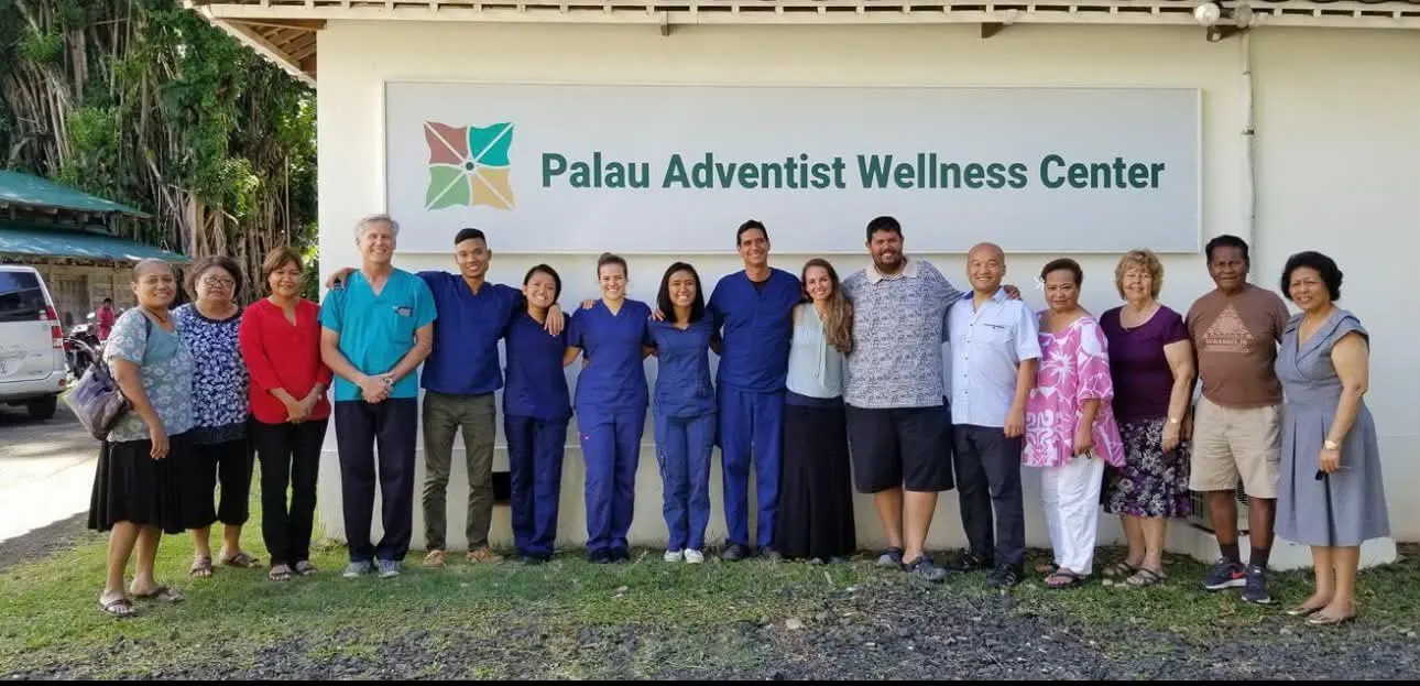 Clinic staff and volunteers standing in a line in front of the Palau Adventist Wellness Center sign