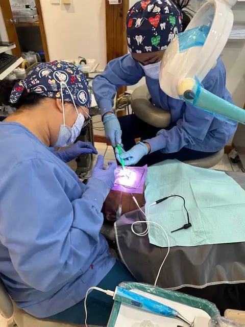 Two dental clinicians in caps and gowns treating a patient with a curing light