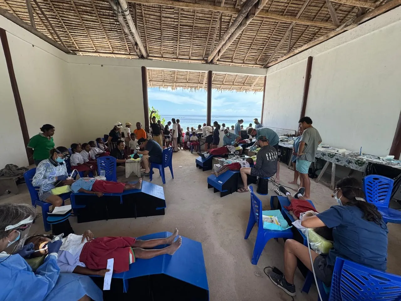 Volunteer dentists treating schoolchildren on portable dental beds in a thatched hall open to the sea, with a crowd waiting