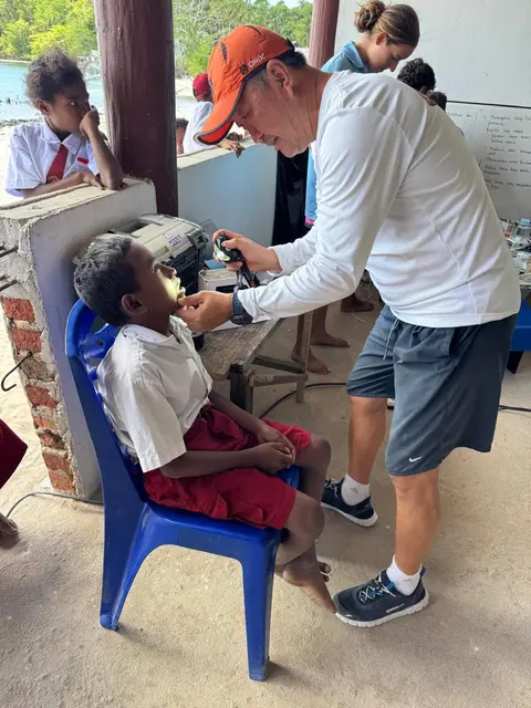 A volunteer dentist in an orange cap examining a schoolboy’s mouth with a light