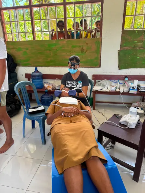 A dentist cleaning a student’s teeth as children peek through the window bars