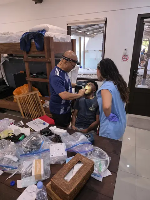 A dentist with a headlamp and a young volunteer treating a seated patient beside a table of supplies