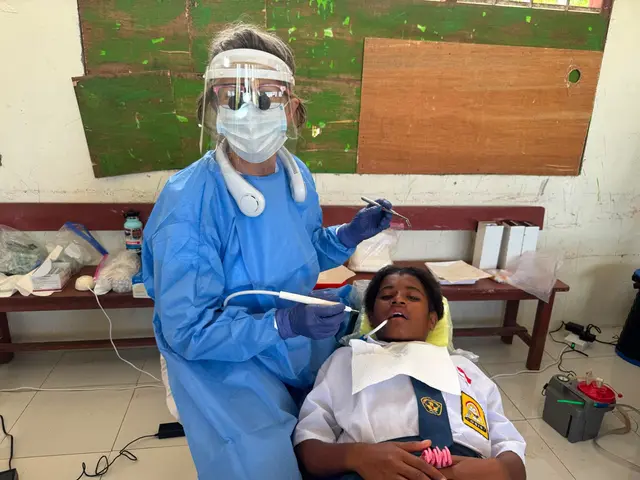 A dentist in a face shield and gown beside a schoolgirl in the dental chair