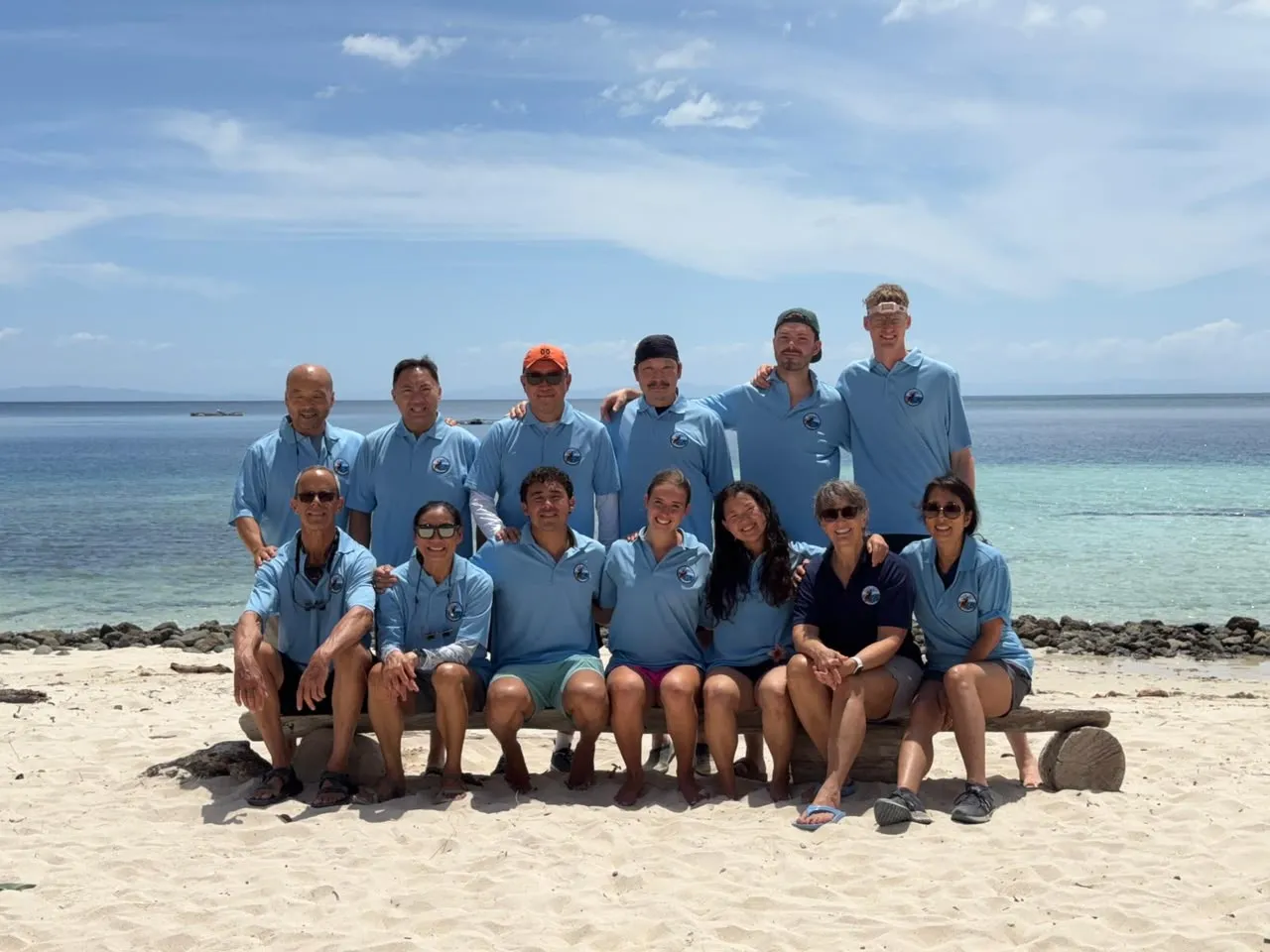 The Raja Ampat mission team in matching light-blue shirts, sitting and standing together on a white sand beach