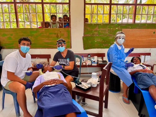 Volunteer dentists treating two schoolgirls on portable dental beds while children watch through the classroom window